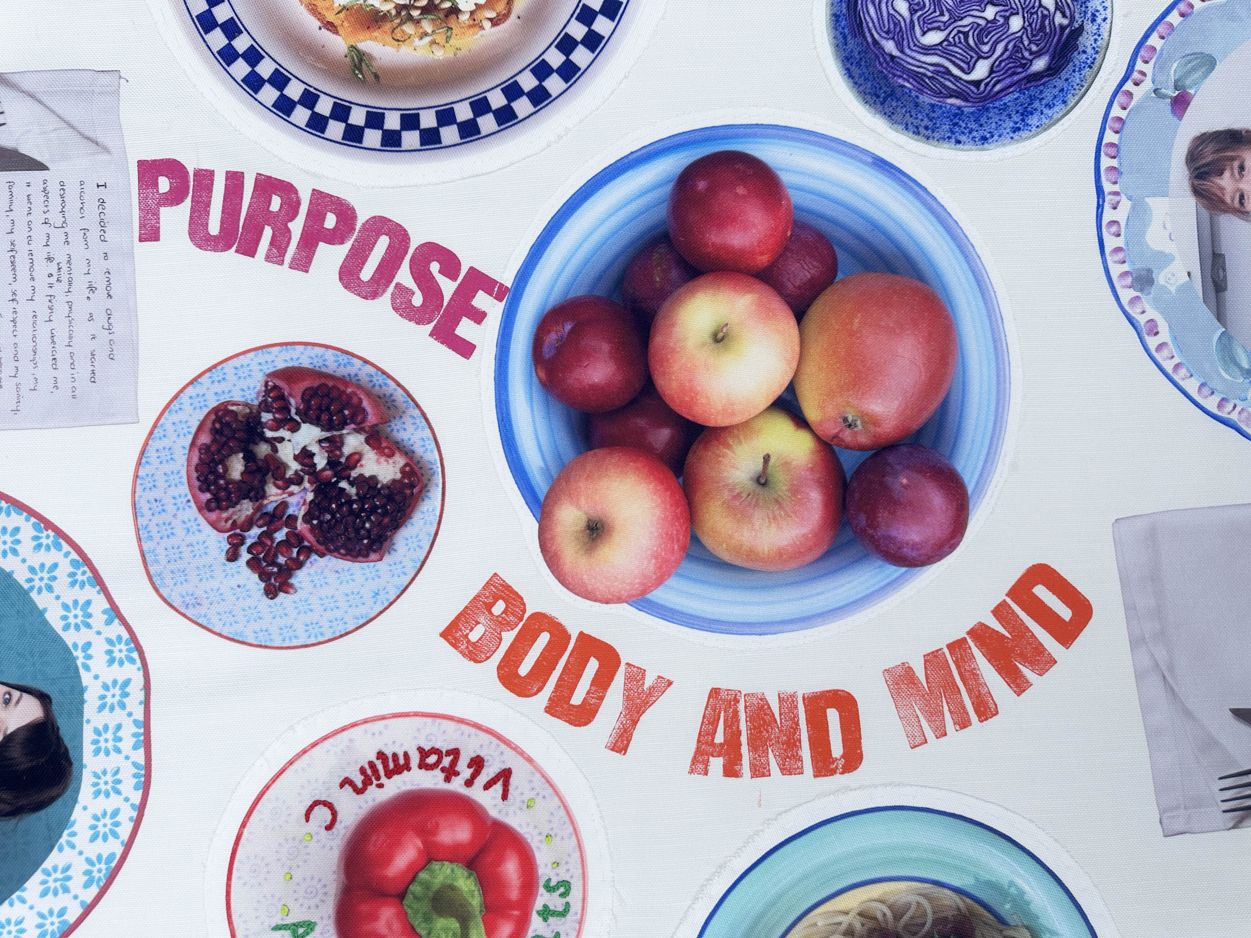 Blue bowl of red and yellow apples surrounded by various plates with food, magazines with "PURPOSE" and "BODY AND MIND" text visible.