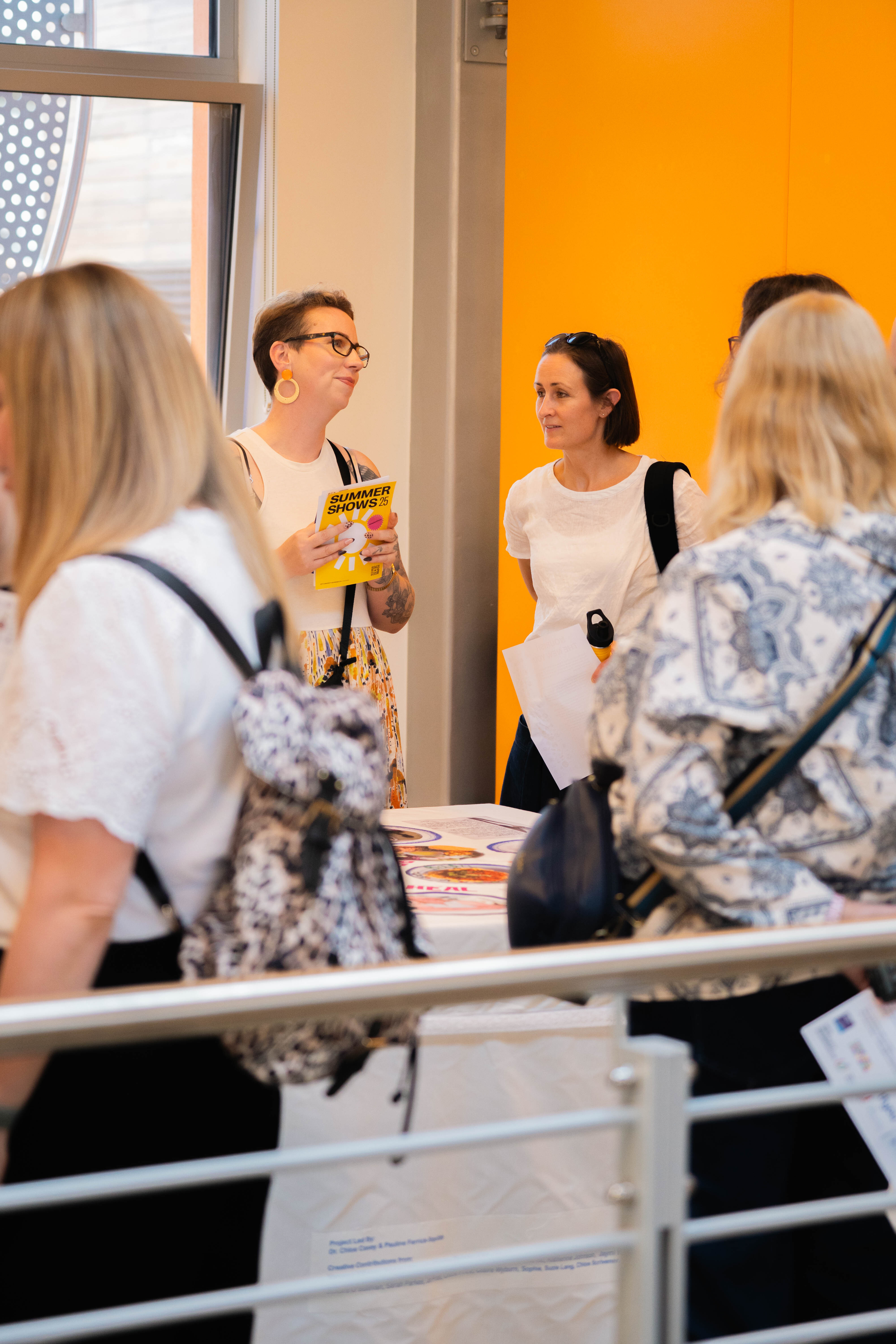 Group of people at indoor event, woman with glasses holding yellow material speaking to others, bright orange wall visible in background.