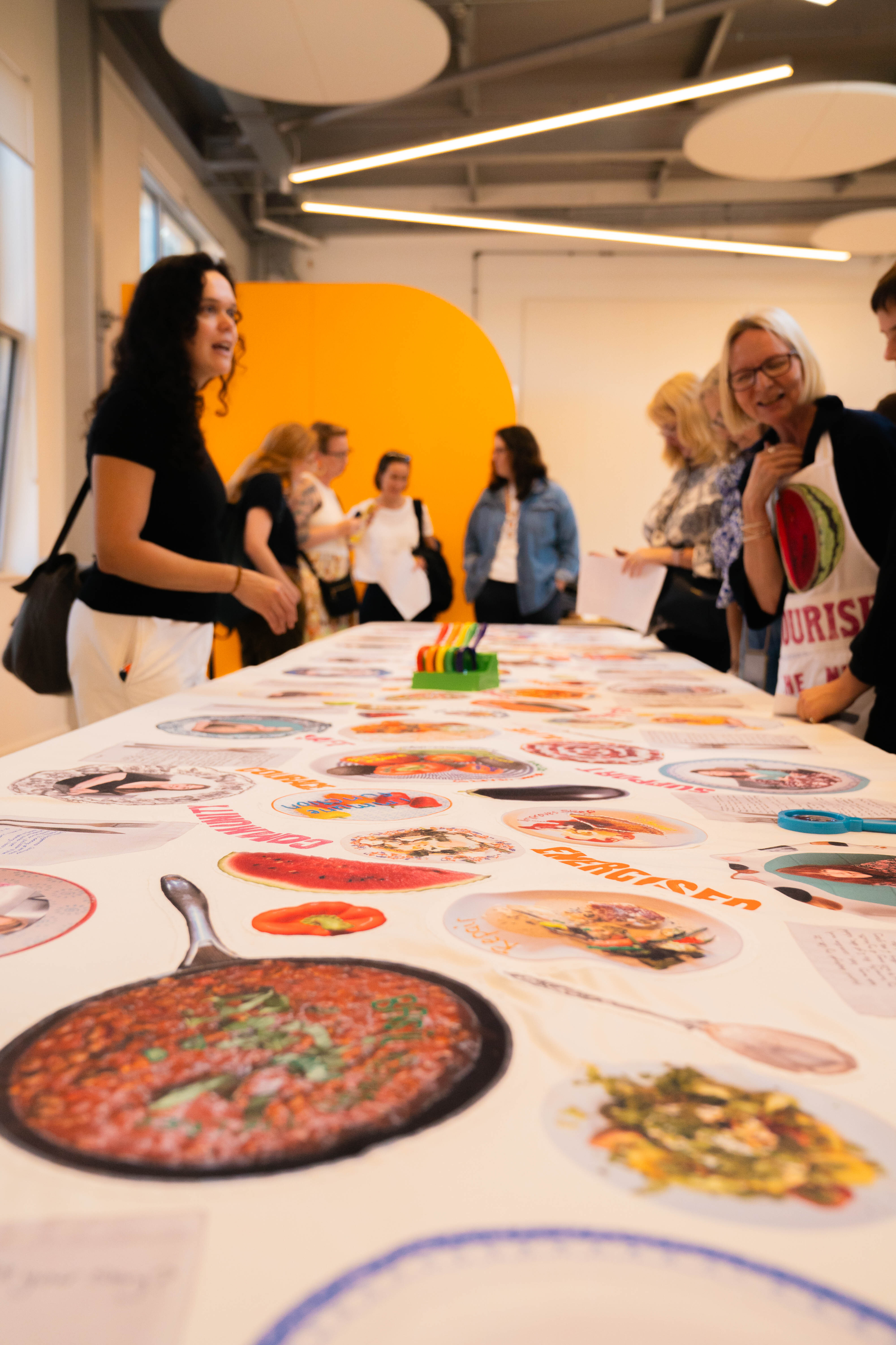 People gathered around white table with colourful food images and circular designs. Orange semicircle on wall, industrial ceiling with round lights.