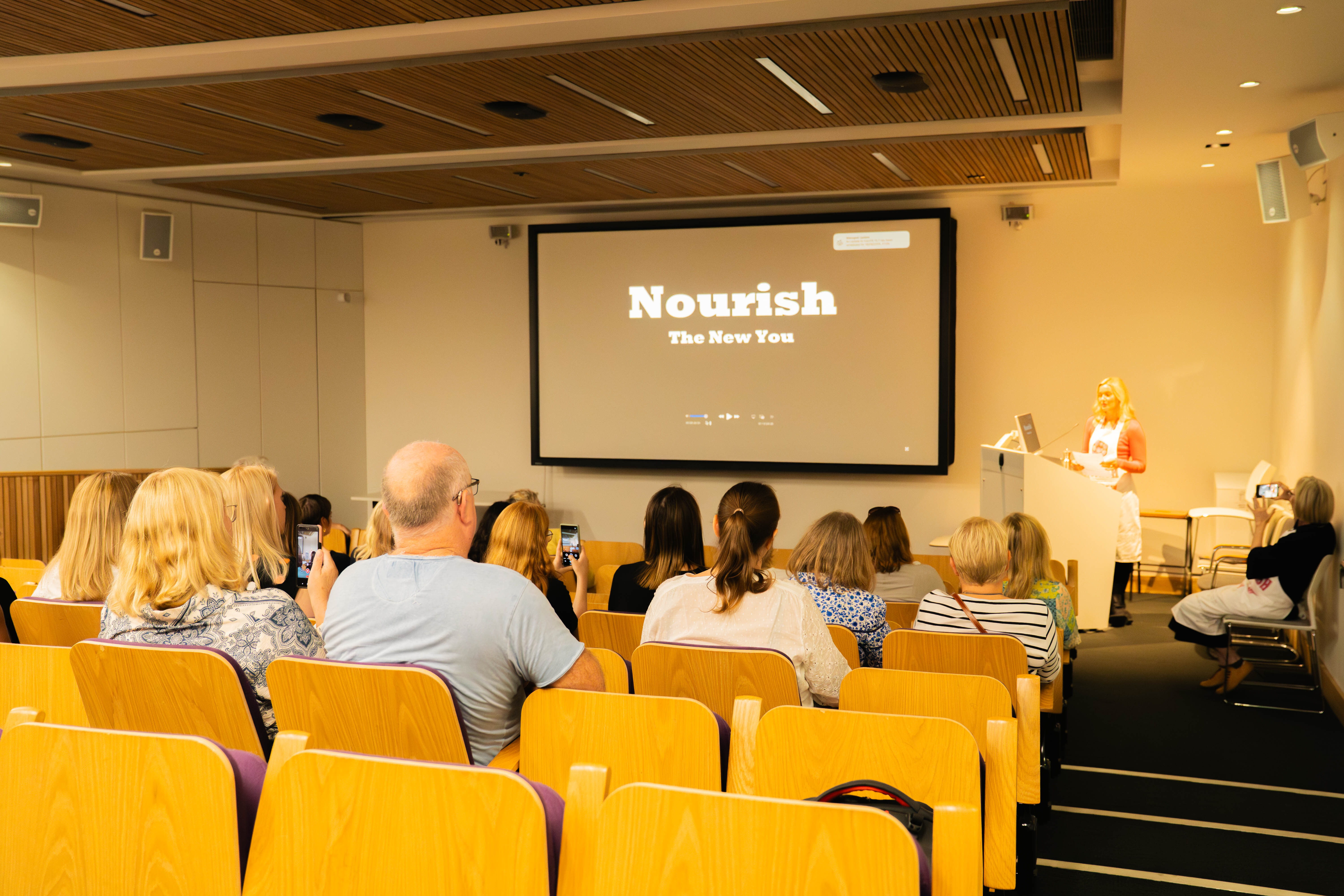 Presentation room with orange chairs, attendees facing screen displaying "Nourish The New You" text, speaker at podium on right.