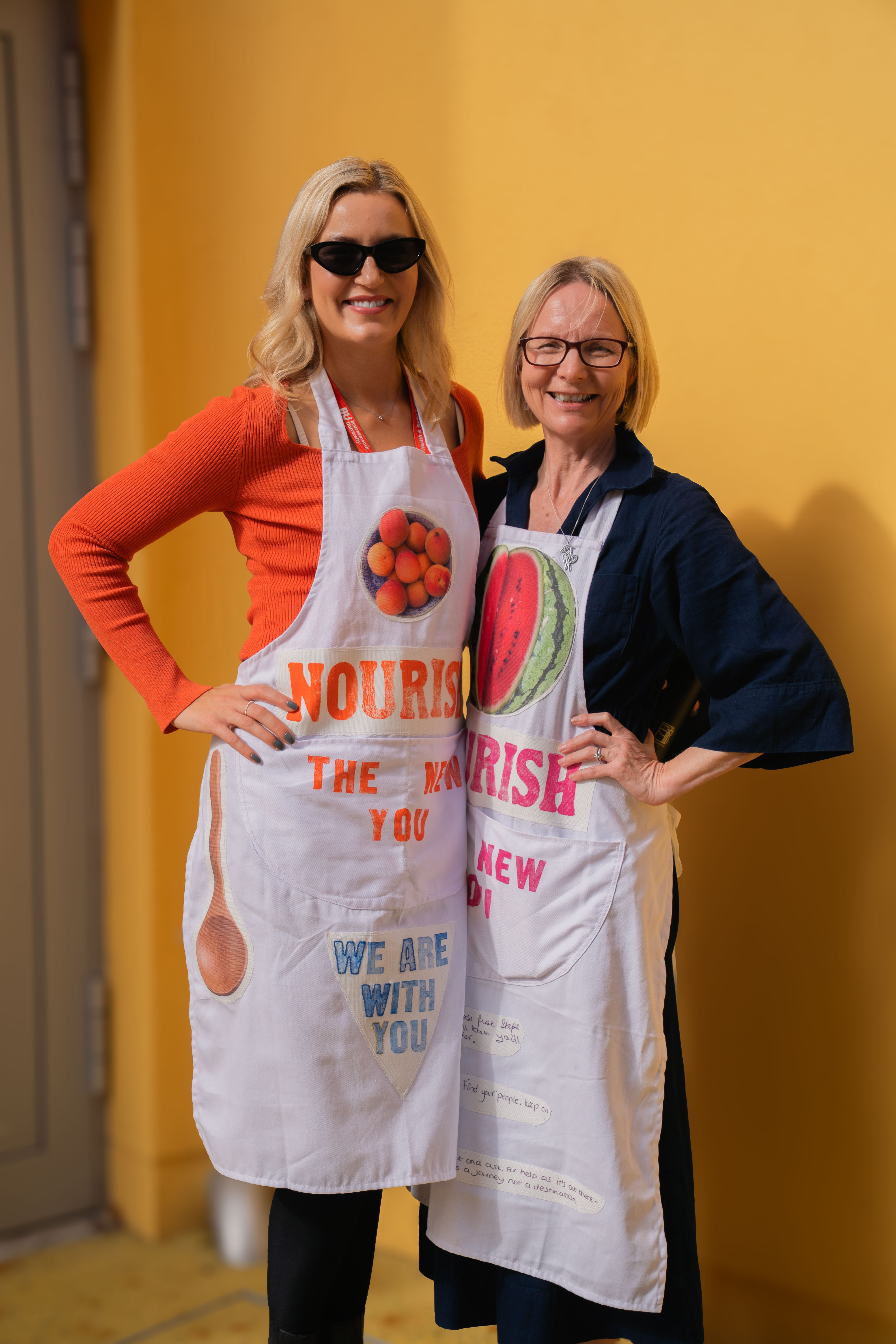 Two women wearing white aprons with fruit graphics and text, standing against yellow wall. Left woman in orange cardigan and sunglasses, right woman in dark jacket with glasses.
