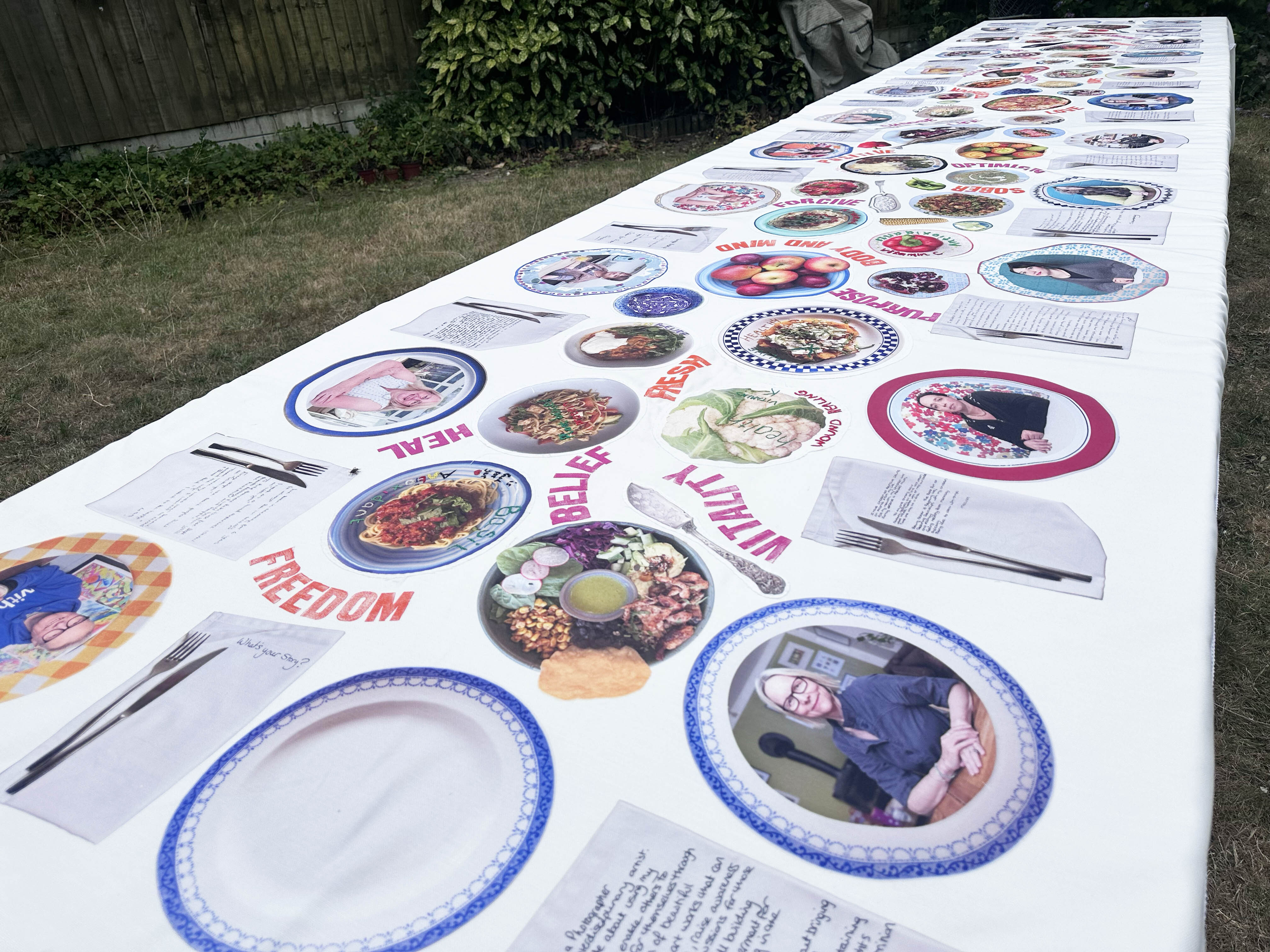 Long white table displaying multiple circular plates with colourful food dishes, empty blue-rimmed plates, and papers arranged in rows outdoors on grass.