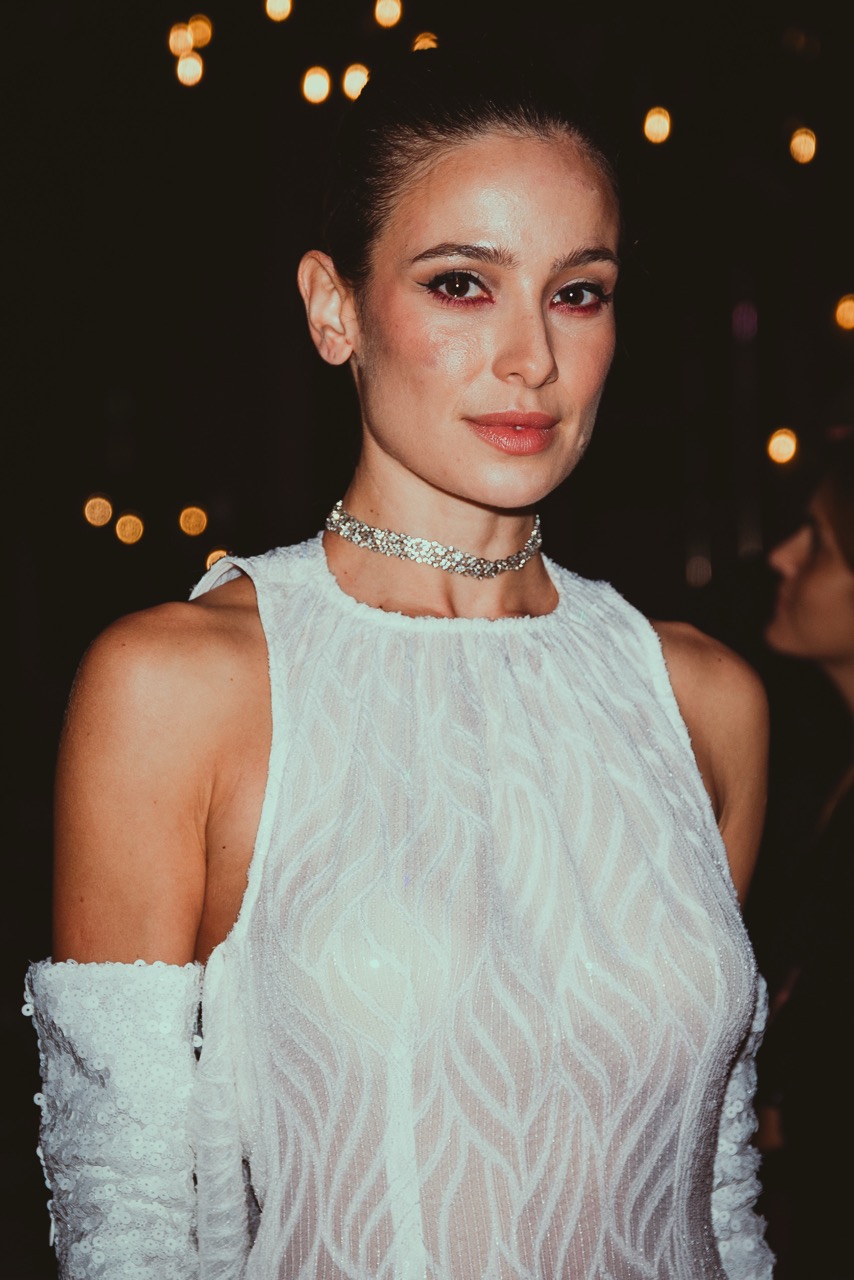 A woman in a white sleeveless dress and matching gloves, wearing a silver choker, poses against a dark background with blurred lights.