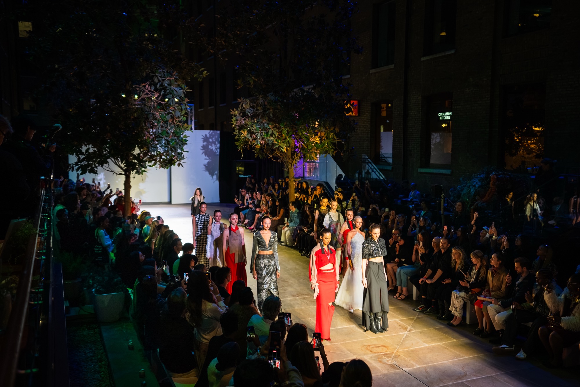 Models walk a runway in evening wear through an outdoor venue, with seated spectators lining both sides.