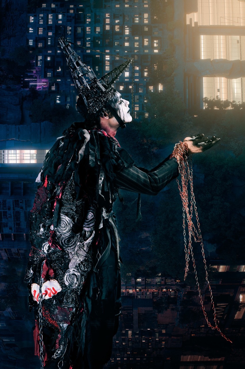 Person in elaborate black costume with spiked headdress holds chain against urban night backdrop with illuminated building windows.