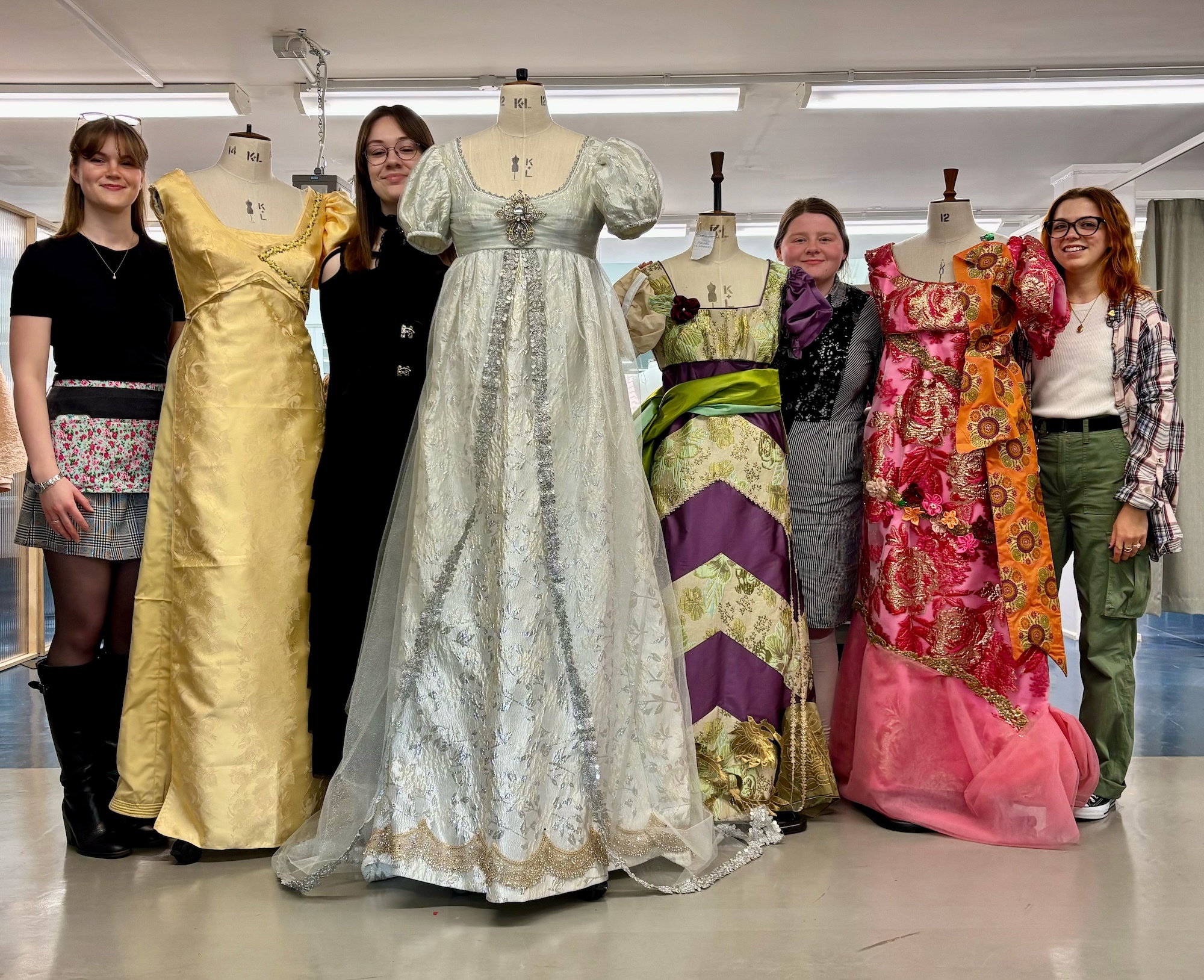 BA (Hons) Costume students O. Wood, L. Stratford, J.L. Donoghue, and J. Little standing next to colourful Regency-style dresses displayed on mannequins.