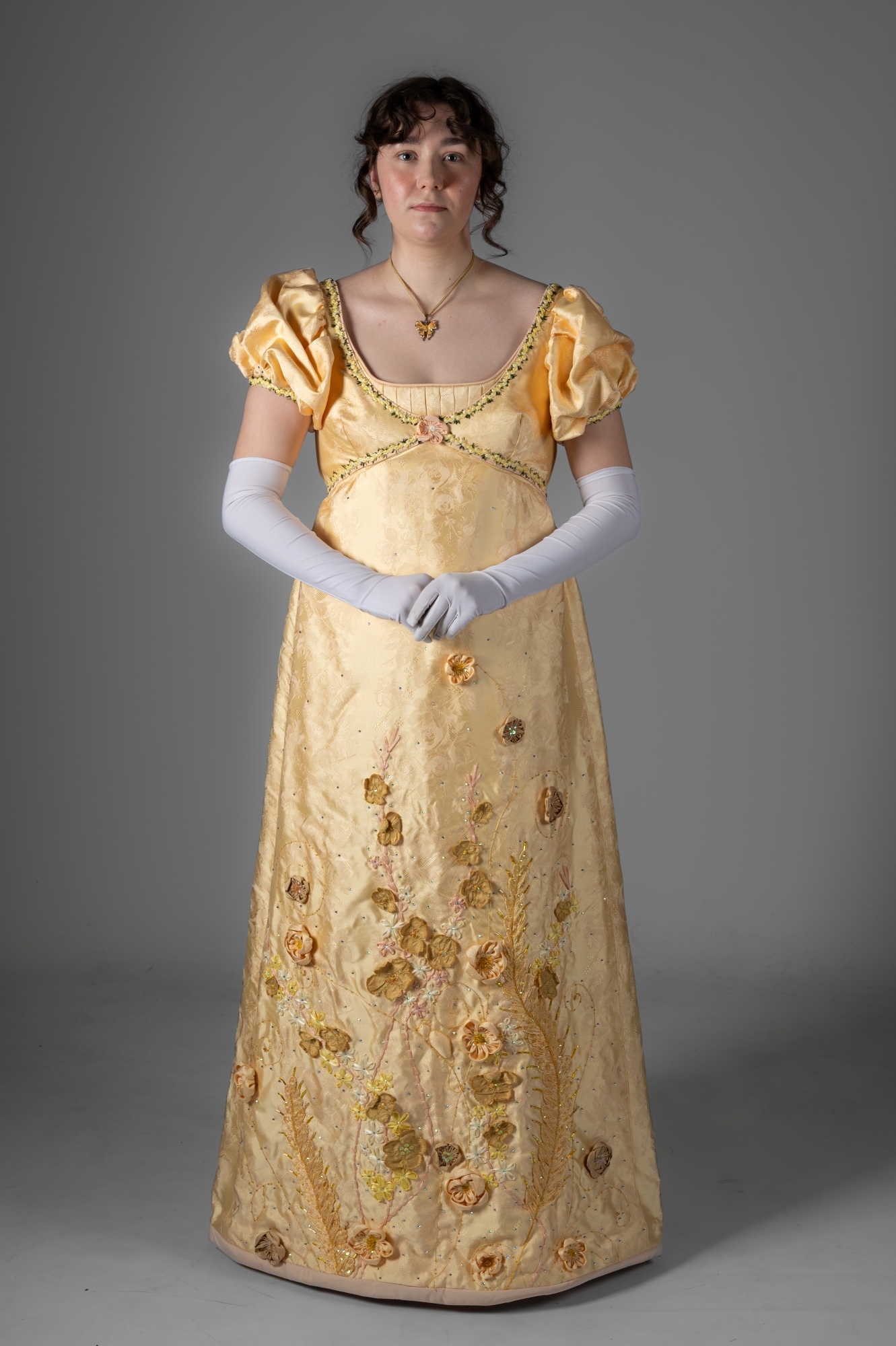 Woman in golden Regency-era gown with puffed sleeves, floral embroidery, and white gloves against grey background.