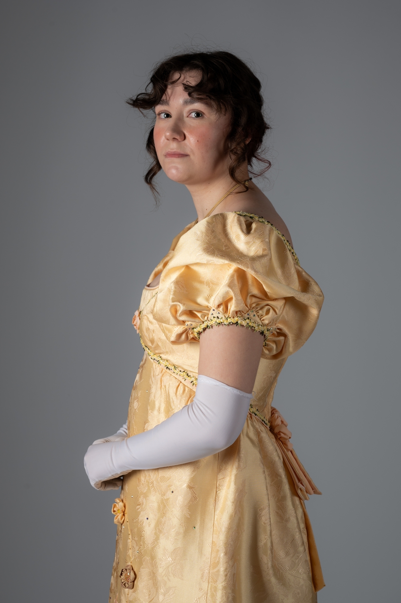 Woman in golden Regency-era gown with puffed sleeves and empire waist, wearing white gloves, against grey background.