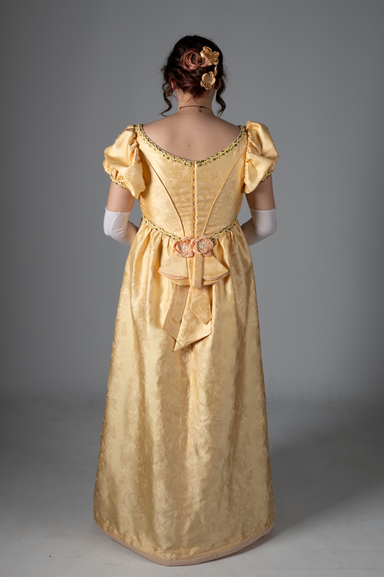 Woman in yellow silk Regency-era gown with puffed sleeves, lace-up back, and decorative chain. Hair in updo with flowers.