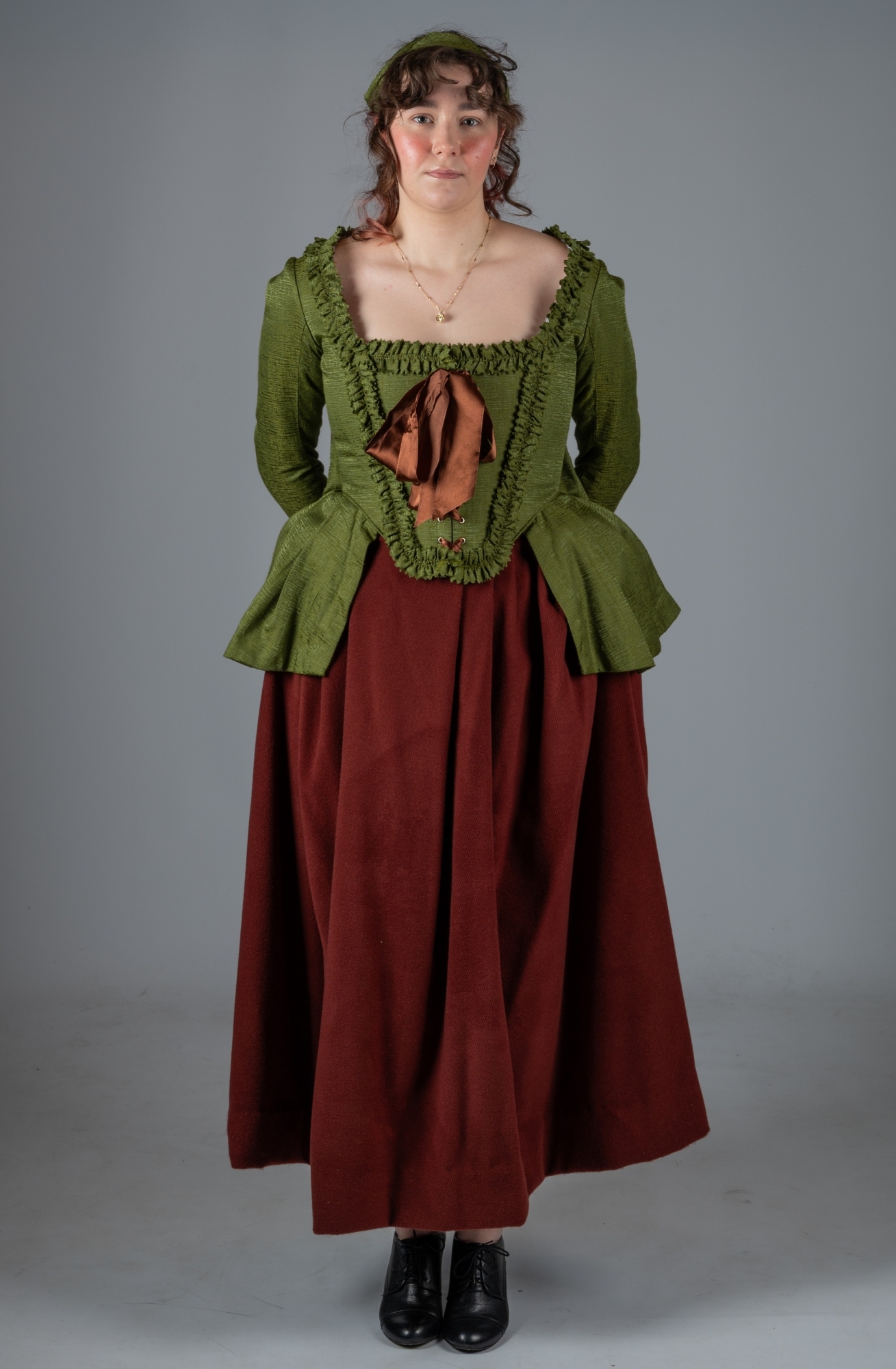 Woman in period costume with green ruffled bodice over burgundy dress, black boots, curly hair, grey background.