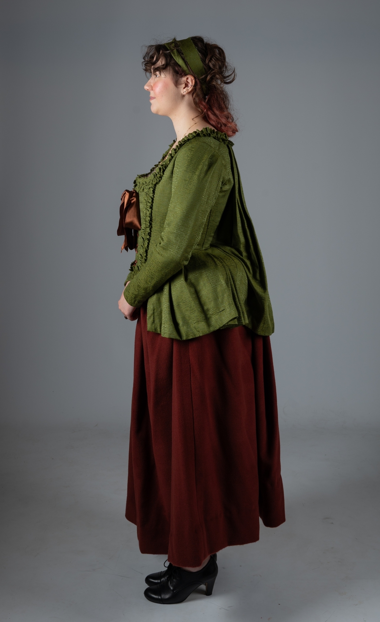 Woman in profile wearing olive green jacket with ruffled trim, burgundy skirt, green hair ribbon, and black shoes against grey backdrop.