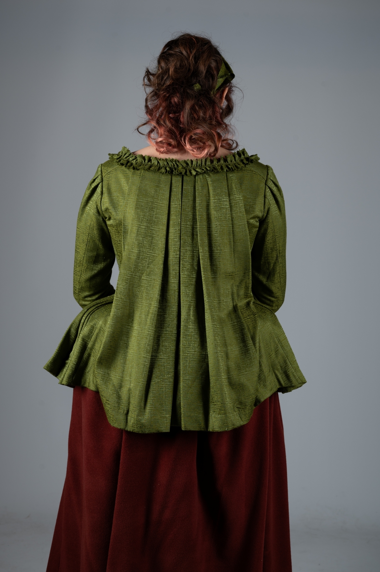 Woman with curly brown hair facing away, wearing green fitted jacket with ruffled collar and burgundy skirt against grey background.