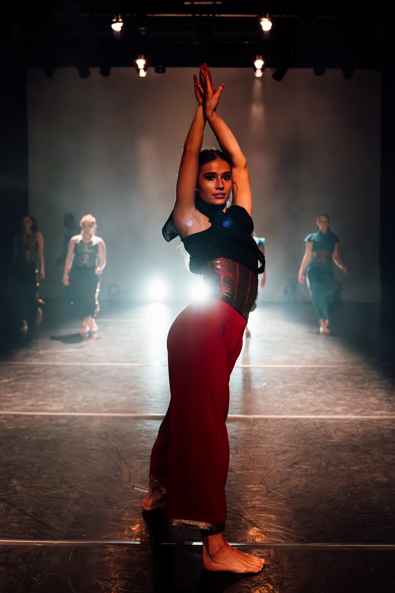 Woman in black top and red trousers dancing on stage with arms raised, other performers visible in background under stage lights.
