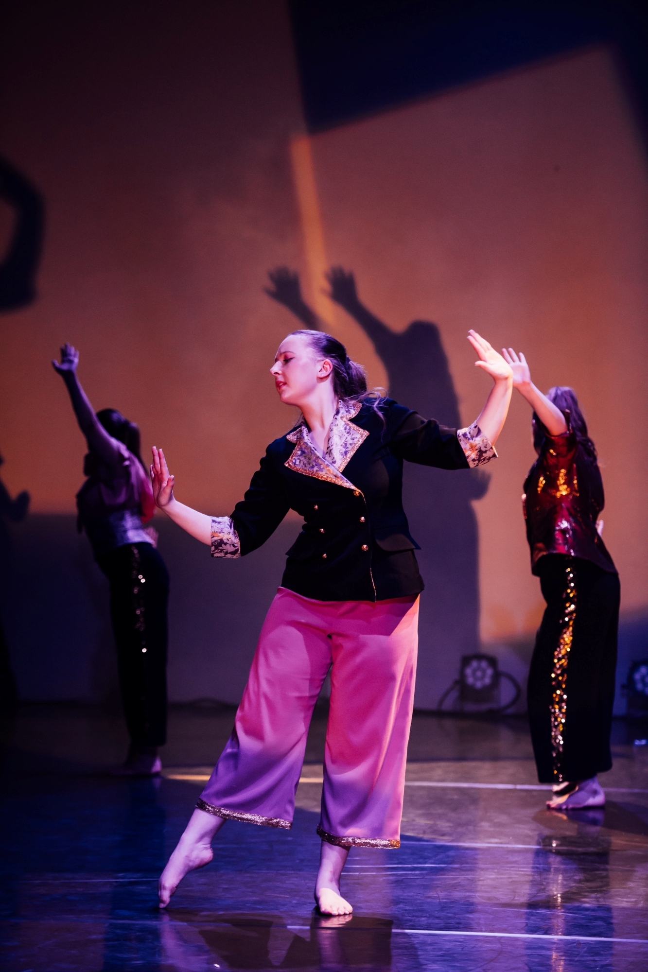 Three dancers on stage with arms raised. Centre performer wears black top and pink trousers. Warm orange and purple stage lighting creates dramatic shadows on backdrop.