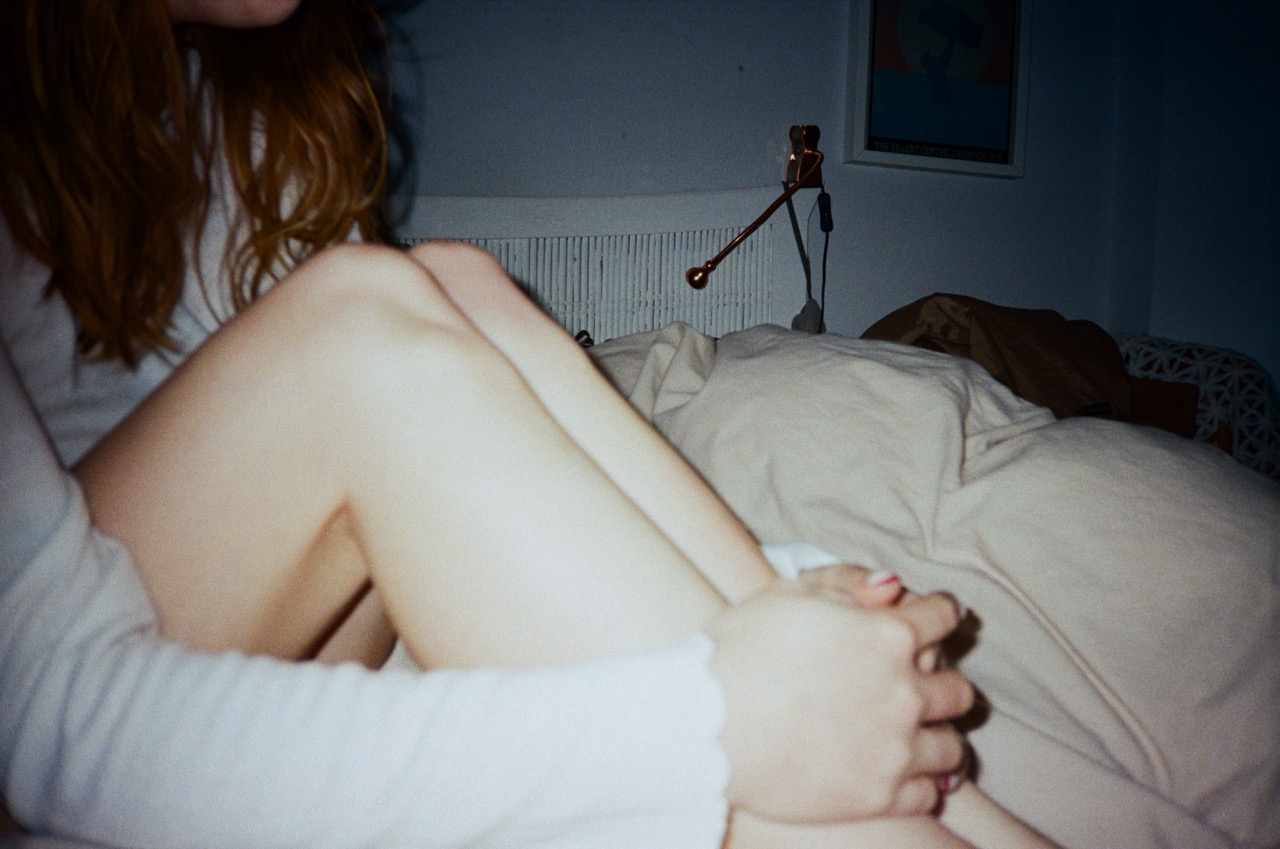 A close-up of a person's legs, with bare knees crossed and hands clasped around them. The setting features a rumpled bed with light-coloured bedding, and a softly lit background with a wall-mounted lamp and artwork.