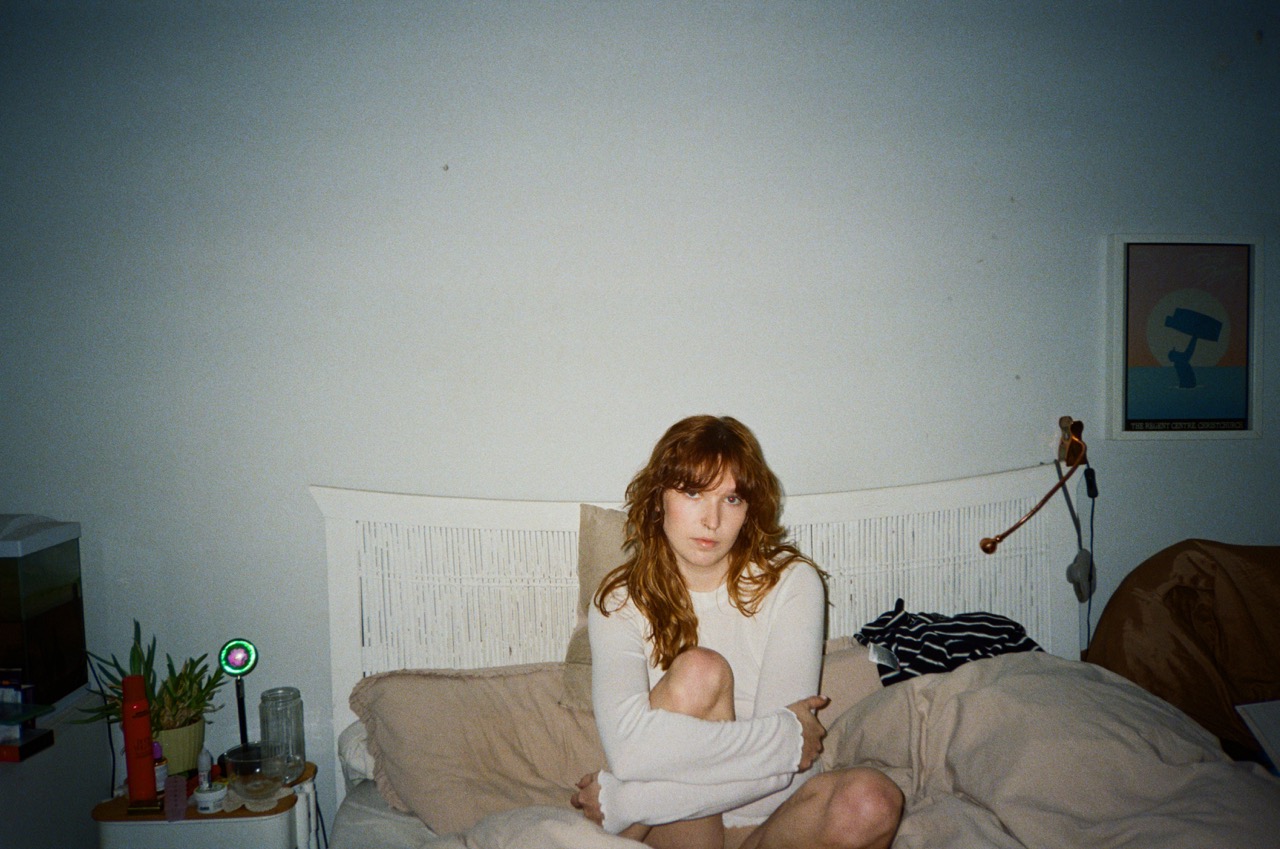 A person with long, wavy red hair sits on a bed covered with beige bedding. The individual is wearing a white long-sleeve top and has their knees drawn to their chest. The background includes a small plant and a framed picture on the wall.