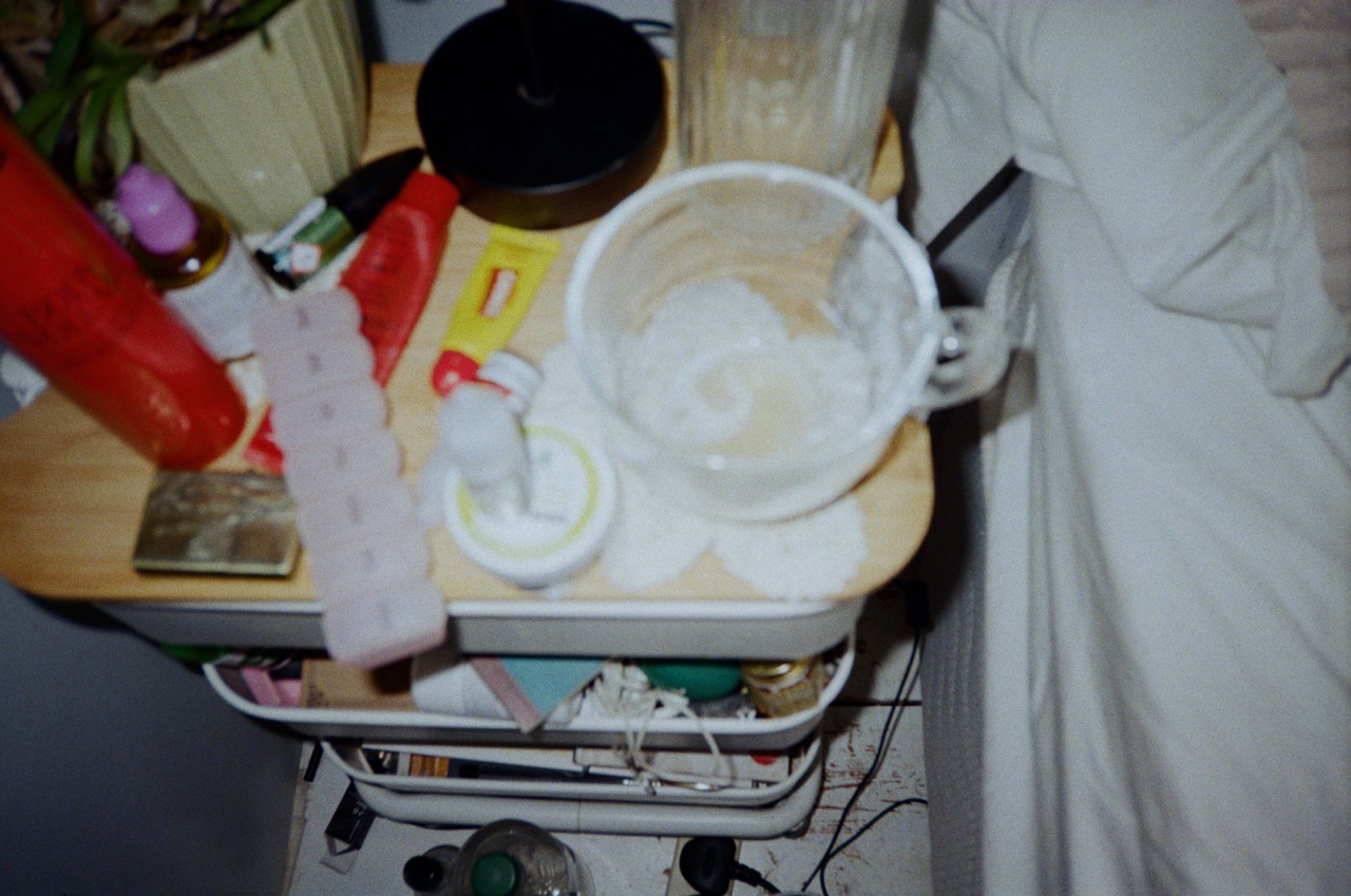 A bedside table cluttered with various items, including a bottle of lotion, a glass of water, a pink pill organizer, and plants. An empty plate and a small jar are also visible on the surface.