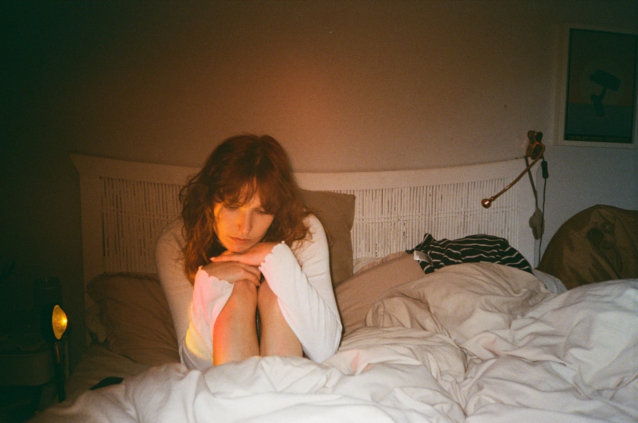 A young woman with shoulder-length brown hair sits on a bed, hugging her knees. The bed is covered with white bedding, a table lamp emits warm light, and there is a framed picture on the wall in the background.