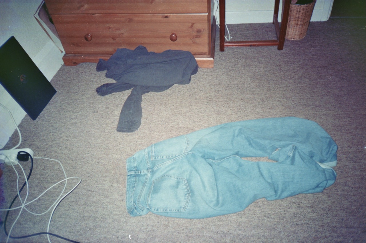 A pair of blue denim jeans laid flat on a light-coloured carpet, next to a grey sweatshirt draped partially on the floor. A wooden chest of drawers is visible in the background, along with a small wooden shelf. A black device and white cables are also seen.