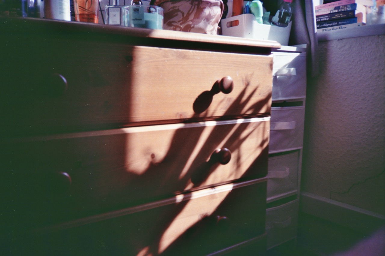 A wooden dresser with three drawers and round handles, illuminated by natural light, casts a shadow on the floor. In the background, a clear plastic storage unit and various containers are visible.