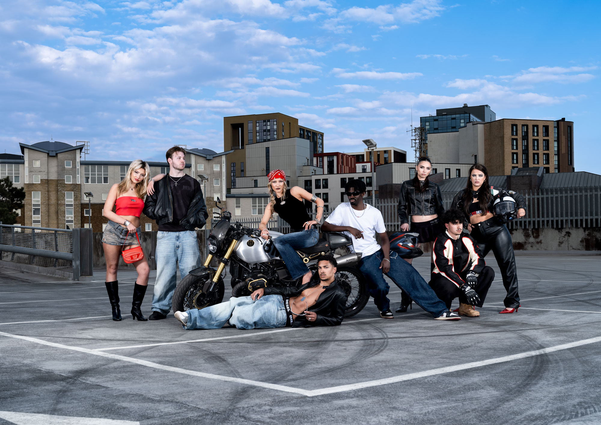A group of seven individuals poses on a multi-storey car park rooftop. They are dressed in casual and biker clothing, with some leaning against a motorcycle. The urban background features modern buildings and a partly cloudy sky.