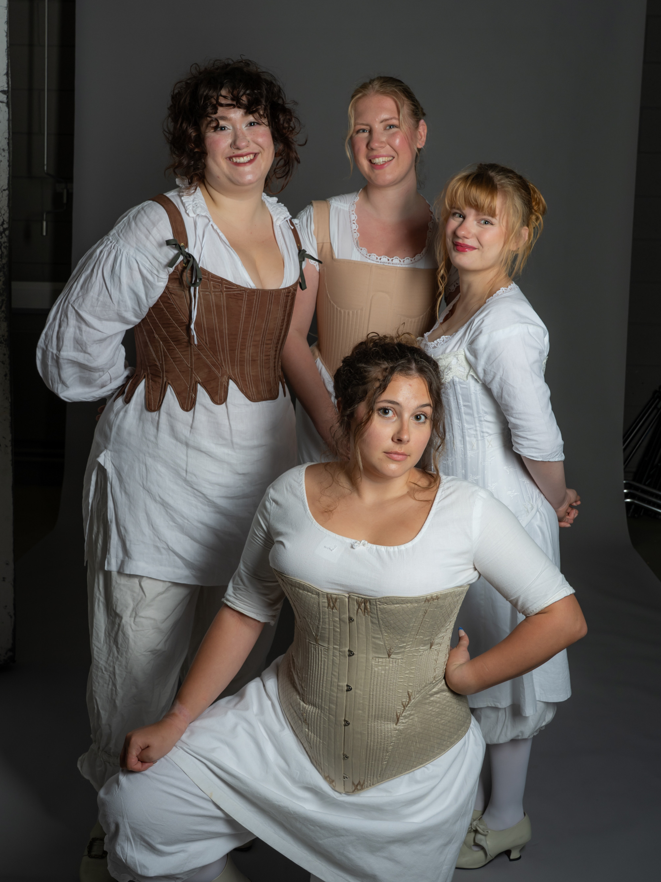 Four women are posed together in vintage-style clothing. Each woman wears a corset and white dresses; they are positioned in a semi-circle, two standing and two kneeling, with a neutral grey backdrop.