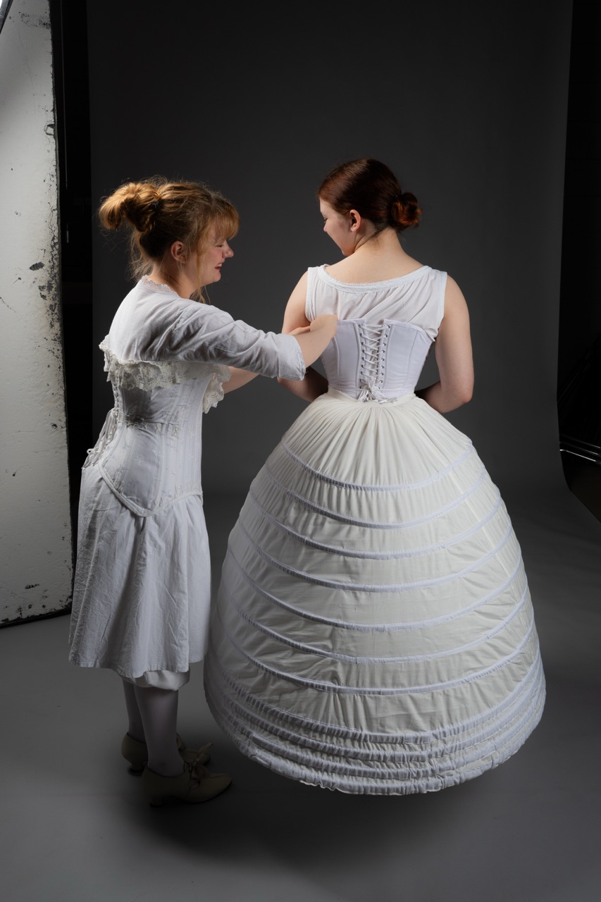 A woman in a white dress is adjusting the corset of another woman wearing a white, full-skirted gown in a studio setting. The background is neutral, enhancing the focus on the attire and the tailoring process.