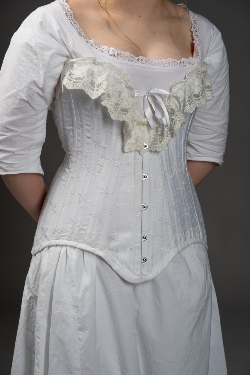 A woman wearing a white corset with lace trim and a ribbon tied at the front, over a plain white dress with long sleeves. The background is neutral, focusing on the attire.