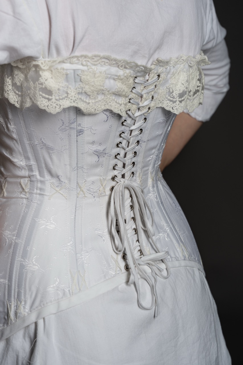 The image shows the back of a white corset with intricate decorative stitching and lace trim along the top. Corset laces are tightly threaded through eyelets, and a tied ribbon at the waist completes the design.