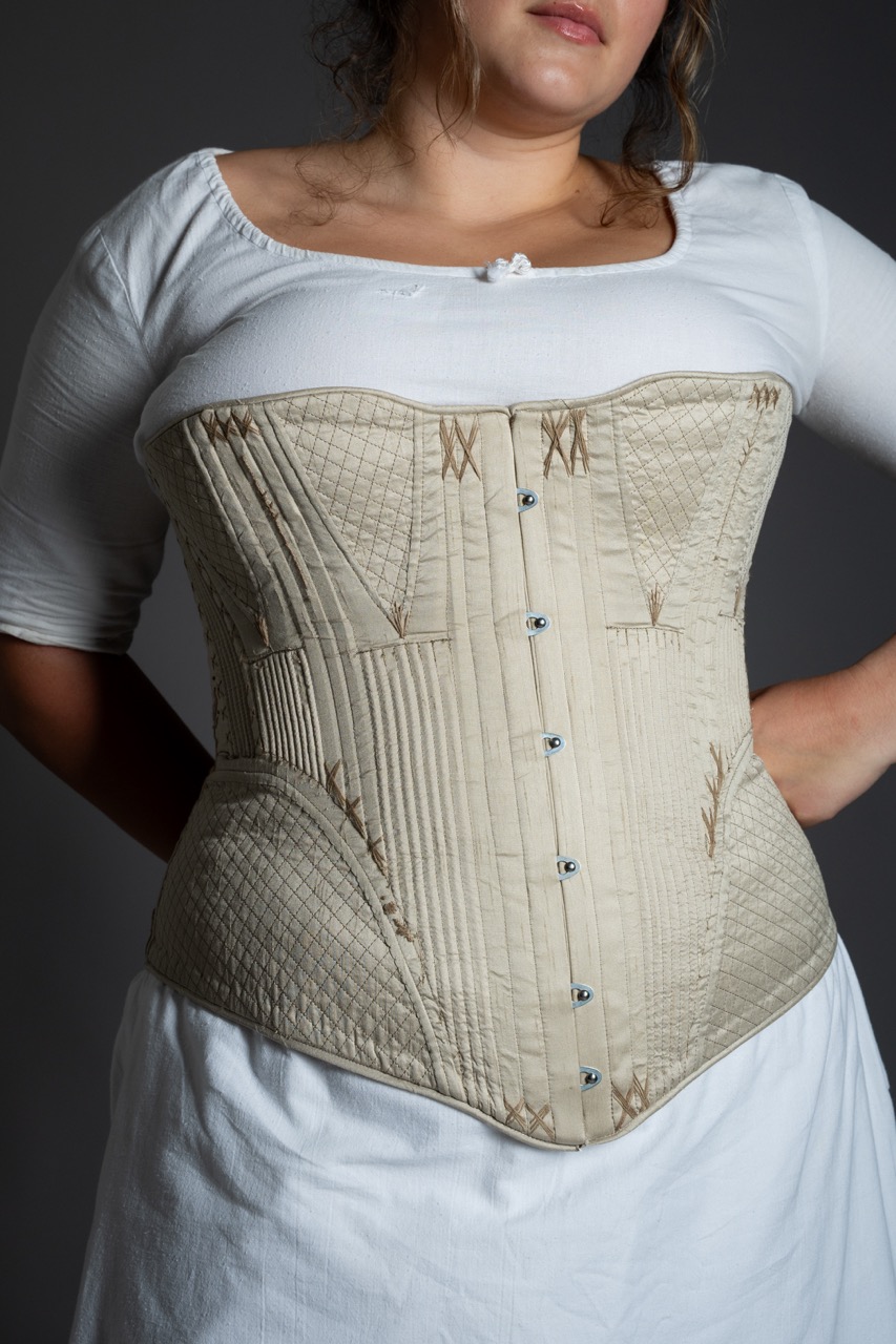 A woman wearing a white dress with a fitted, light beige corset. The corset features vertical stitching and metal fasteners, with decorative elements along the seams. The background is grey, accentuating the details of the corset.