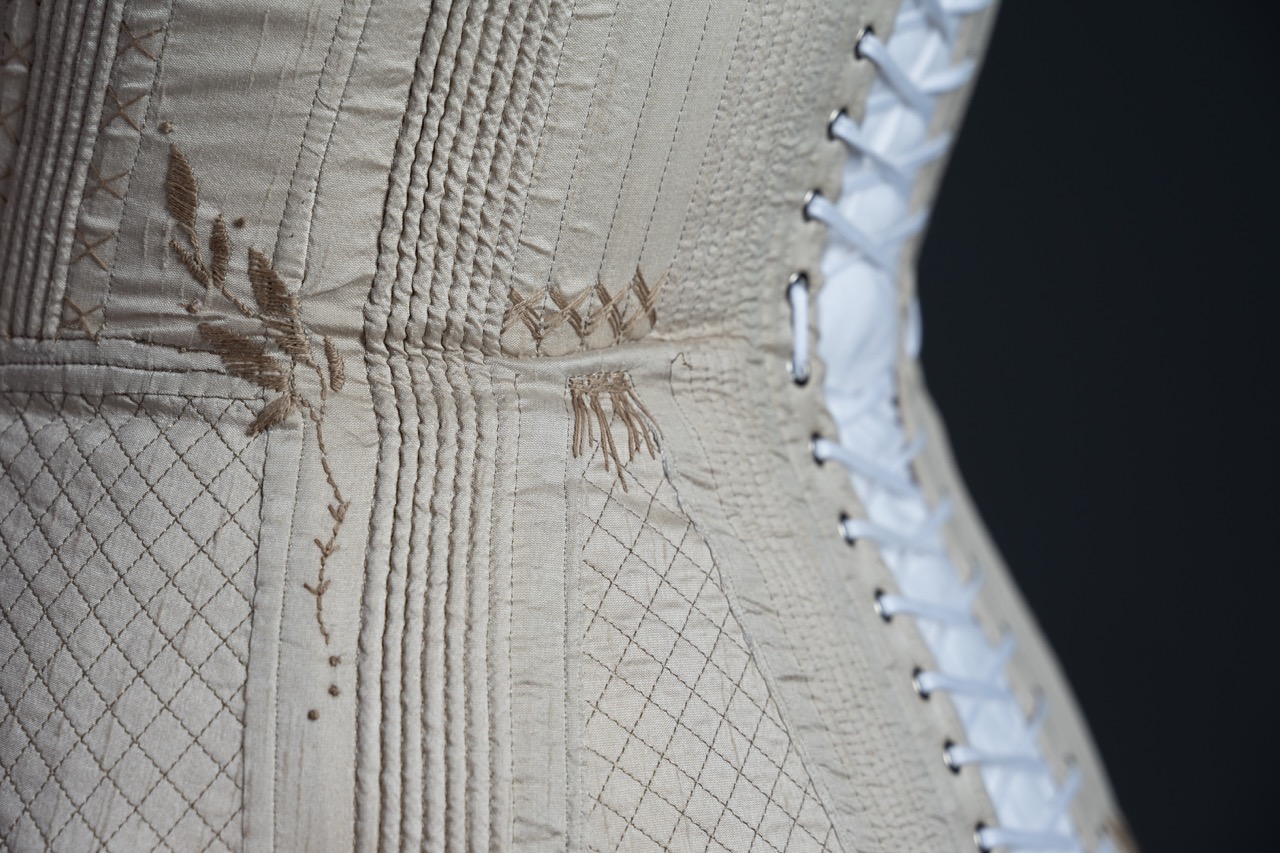 A close-up of a vintage beige corset showcasing intricate vertical stitching and floral embroidery. The corset features a lace-up back in white and is detailed with a diamond-patterned texture across the fabric.