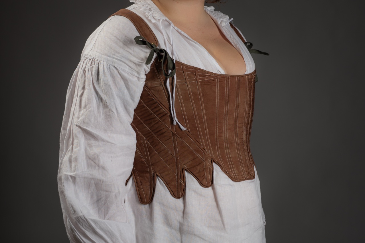 A person wearing a brown leather bodice with a structured design, featuring decorative stitching and laces for fastening. The top is paired with a white long-sleeve shirt, and the background is a plain dark grey.