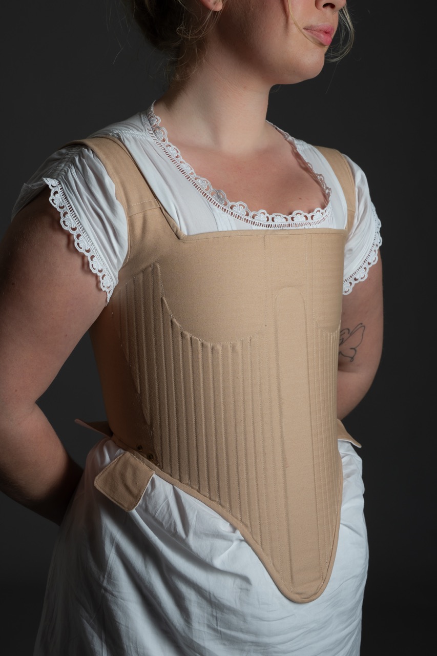 A person wearing a beige corset-style undergarment, featuring vertical ribbing and a structured design, paired with a white blouse that has lace detailing around the neckline. The background is dark, highlighting the garment.