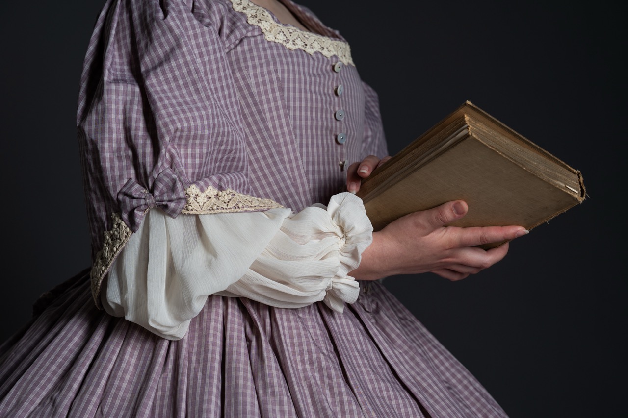 A close-up of a person holding a closed book in their right hand. They are wearing a lavender patterned dress with puffed sleeves, featuring white lace trim on the sleeves.