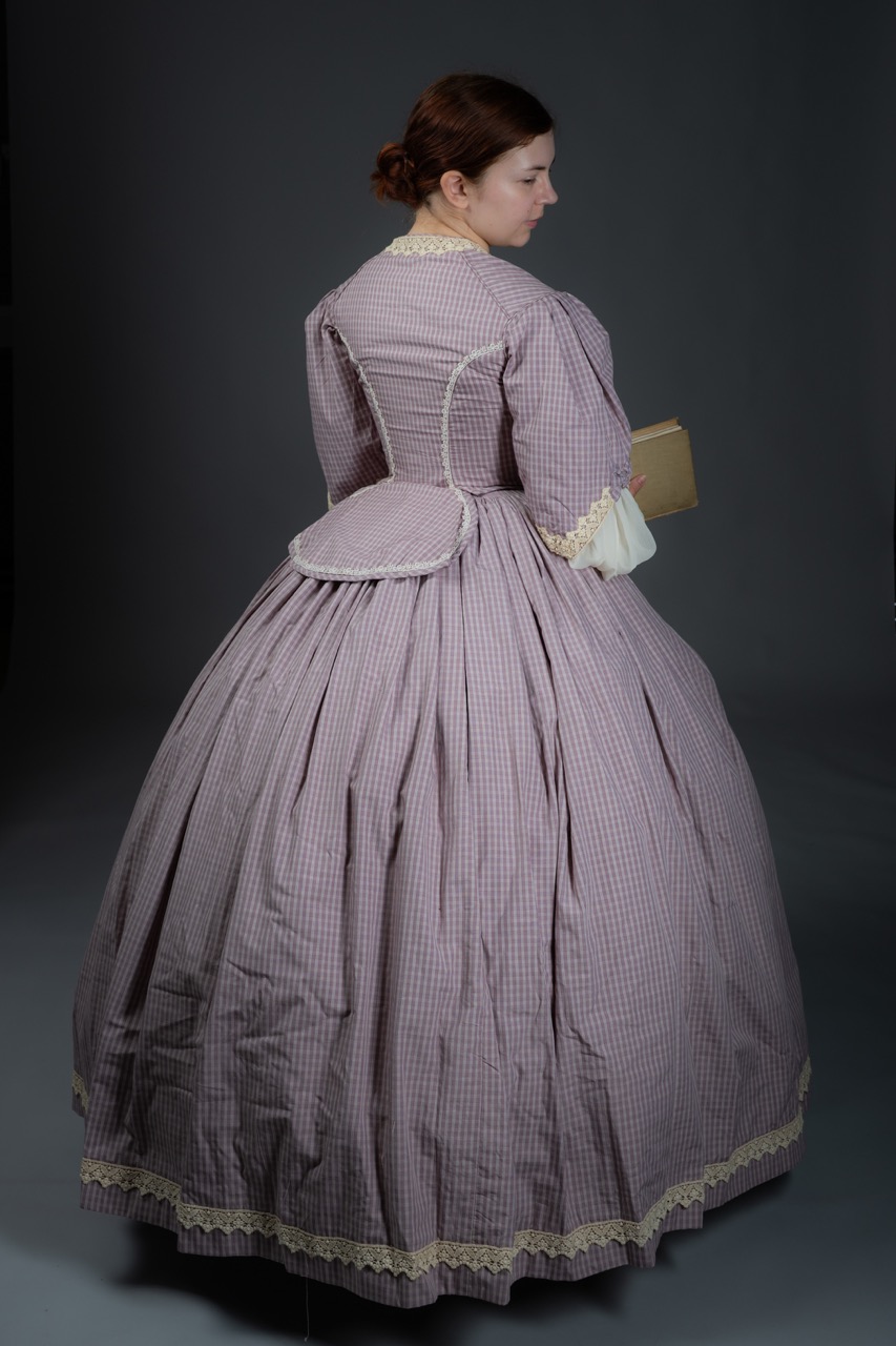 A woman wearing a vintage gown with a full skirt, made of a lavender checkered fabric. The dress features a fitted bodice and a lace trim, with the woman holding a book in her right hand, standing with her back towards the viewer.