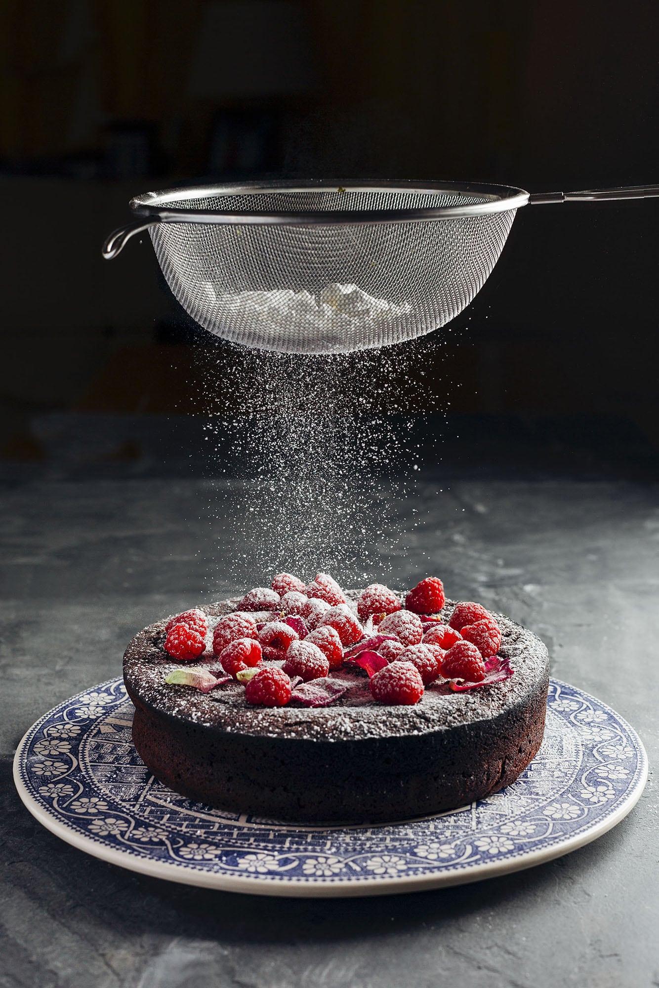 Icing sugar is sifted through a sieve onto a cake sitting on a plate with raspberries on top.