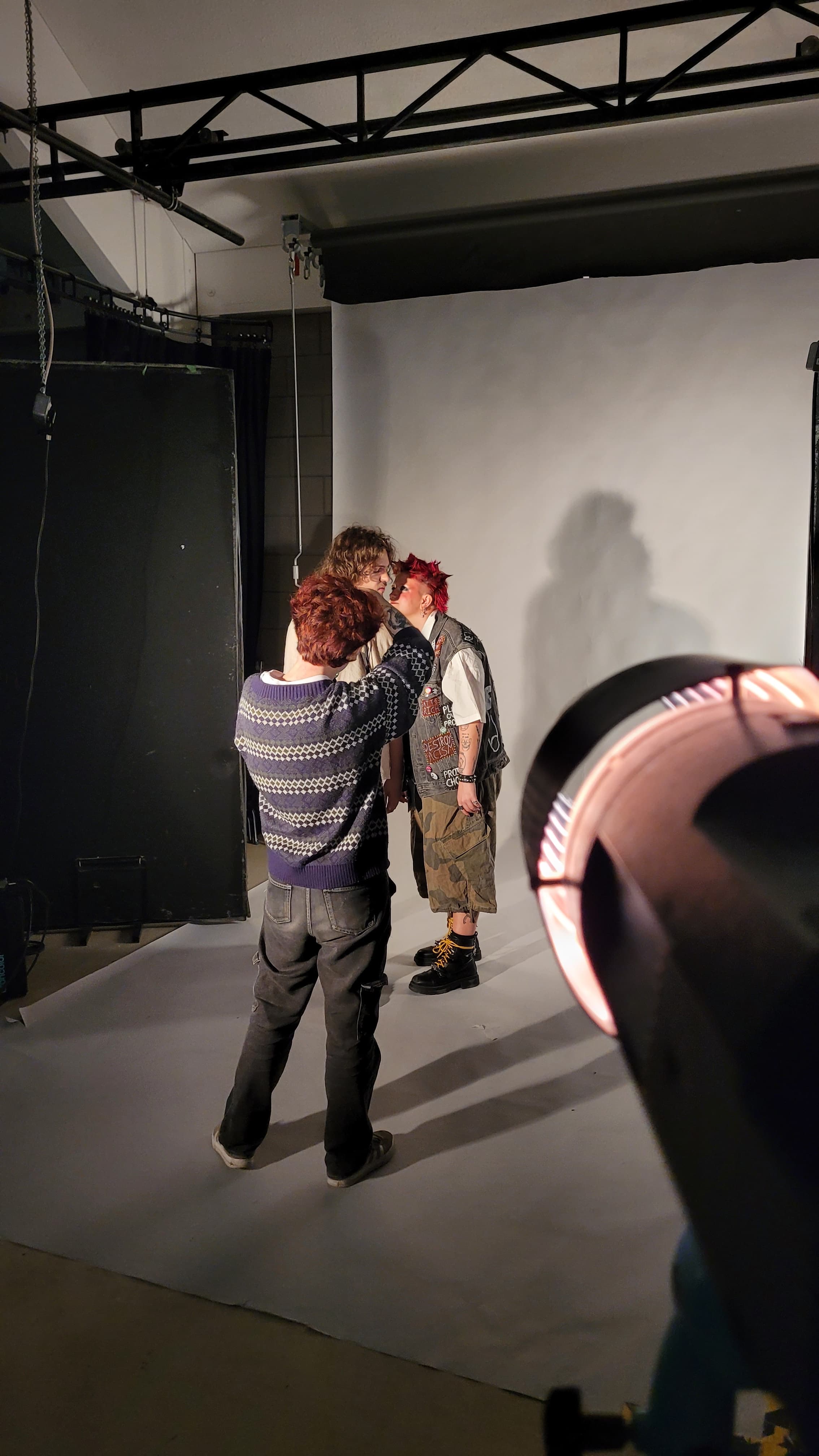 A BA (Hons) Commercial Photography student viewed from behind, standing on a Colorama backdrop in a photography studio taking a photo of two models. A light shines on them from the foreground.