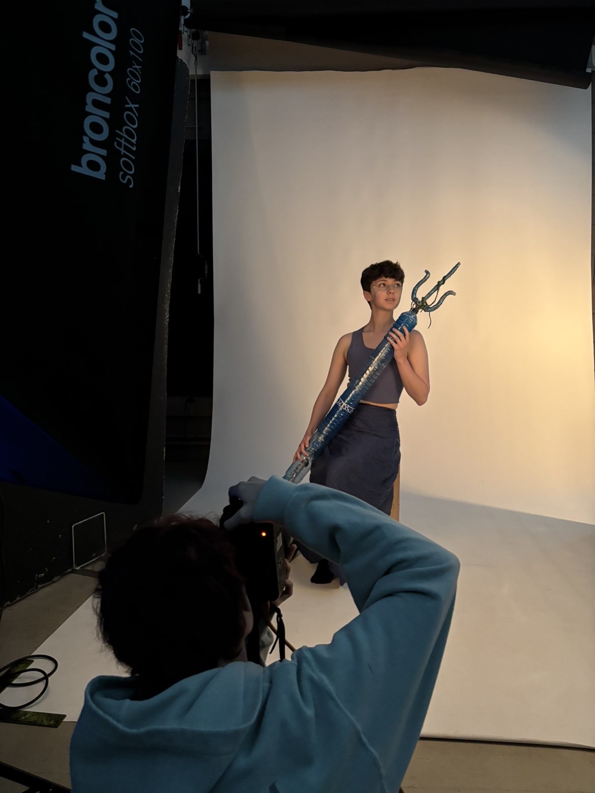 A model stands on a white Colorama backdrop holding a trident-like object, looking off to the side, while a BA (Hons) Commercial Photography crouches in the foreground, taking their photo.