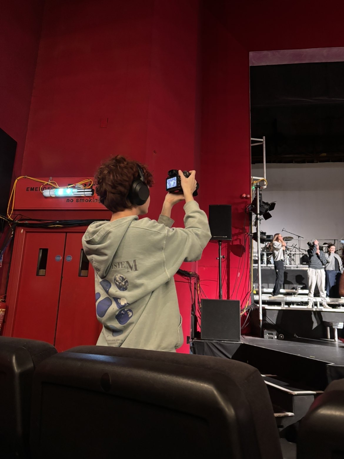 A BA (Hons) Commercial Photography student viewed from behind standing in a theatre space with bright red walls, holding a camera and taking a picture of the stage.