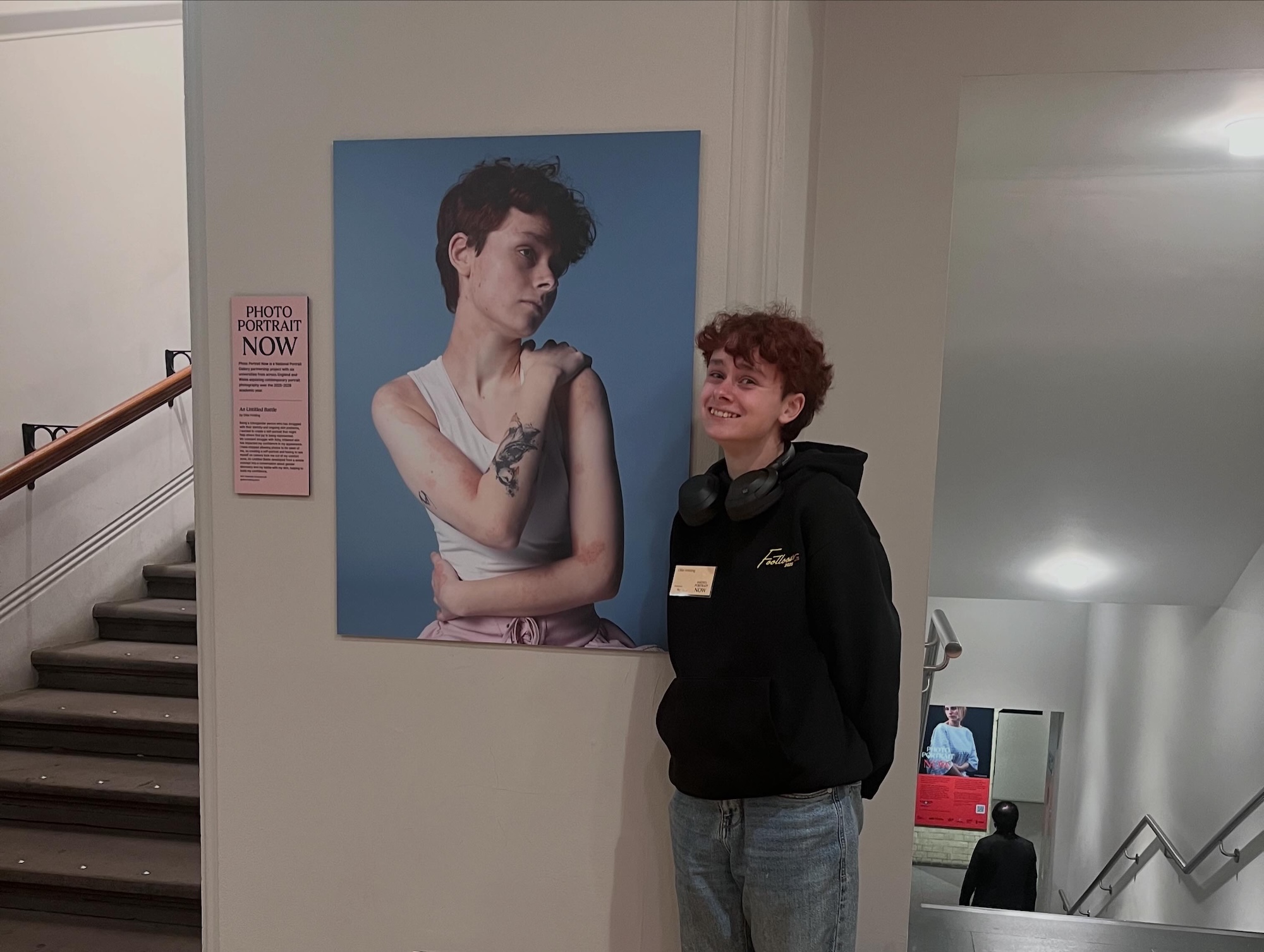 A young person stands beside a large portrait photograph on a gallery wall, with a staircase and steps visible in the background.