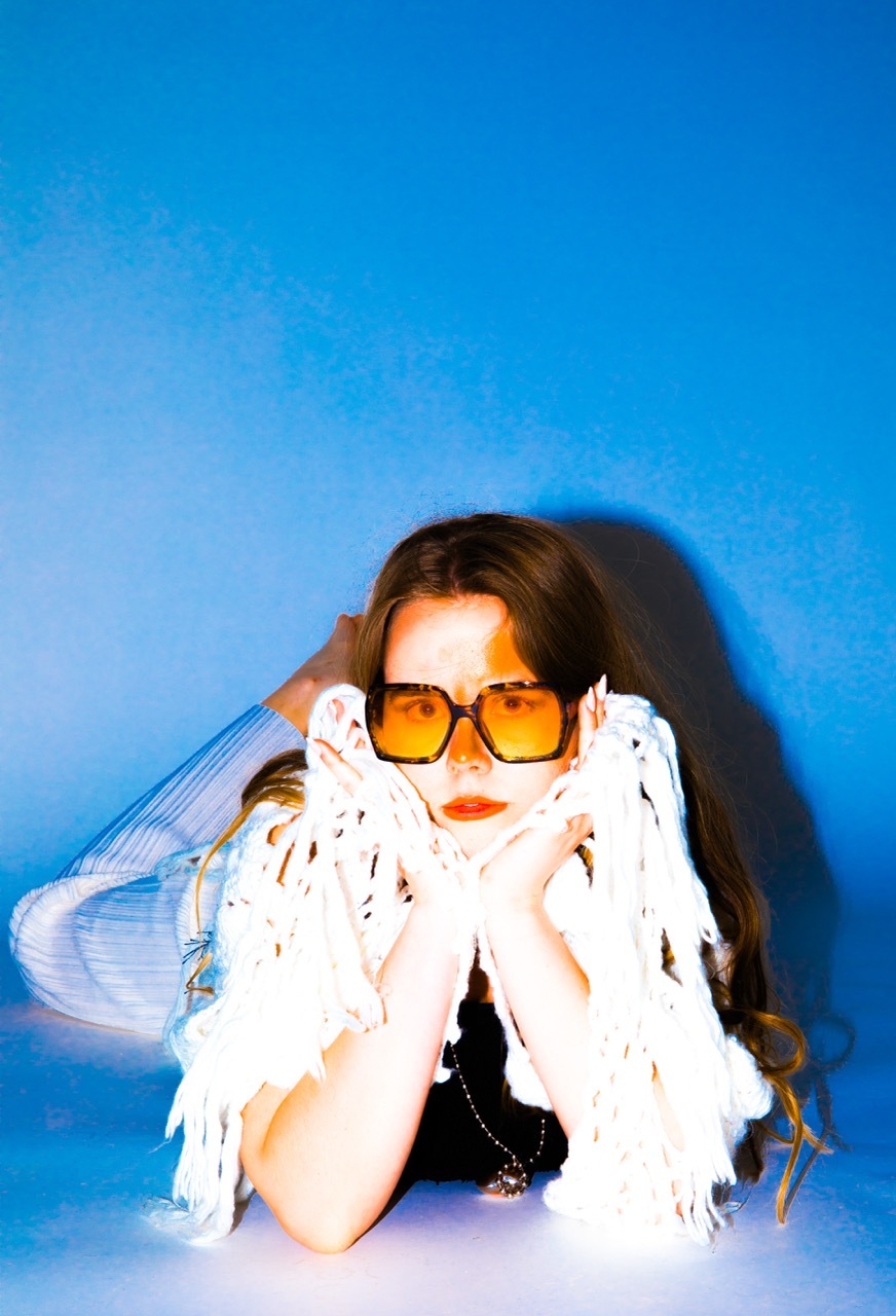 Woman with brown hair wearing orange sunglasses and white feathered garment, hands positioned near face, against bright blue background.