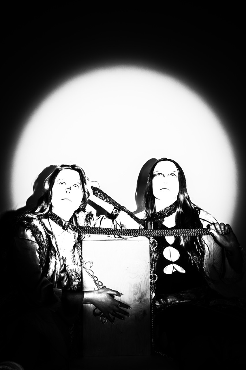 Black and white image of two people in dark clothing with long hair, positioned against a large bright white circular background.