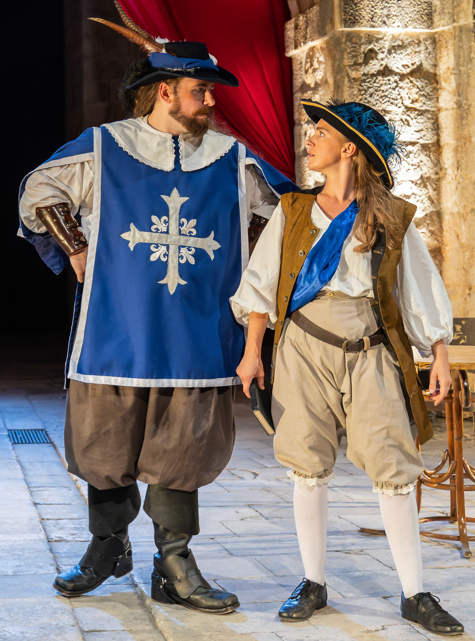 A man dressed in a blue tunic with a white emblem stands next to a woman wearing brown attire and a blue scarf. Both are in historical costumes with period-style shoes, set against a backdrop of red drapery and stone walls.
