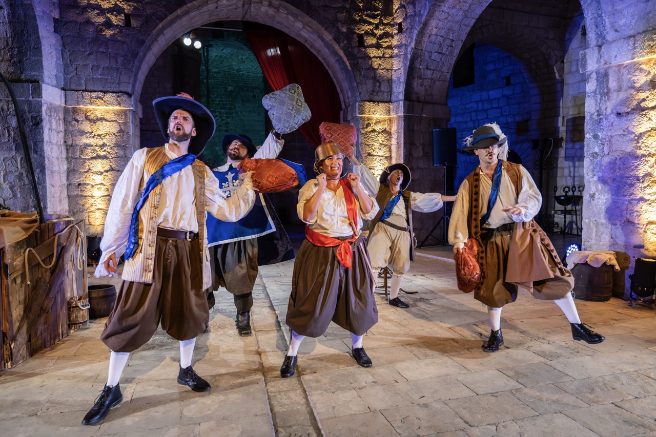 Five performers dressed in historical costumes with ruffled shirts and trousers are performing on a stone stage. They hold various props and display dynamic poses under warm lighting in an arched venue with a rustic ambiance.