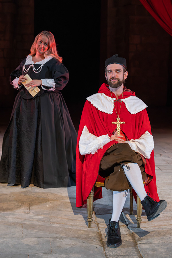 A woman in a black dress with white accessories stands beside a seated man wearing a red cloak and white shirt, holding a cross pendant. They are on a stone stage with a dark background and red drapery.