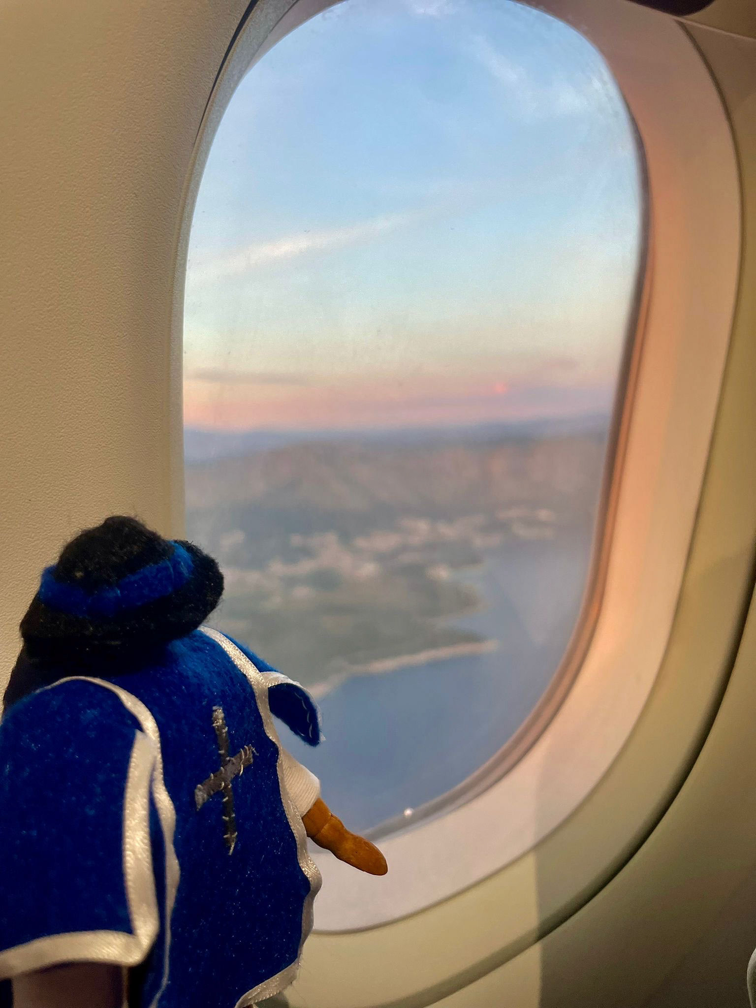A small plush figure dressed in blue clothing sits on an airplane seat next to a window. Outside the window, a scenic view reveals mountains and a lake under a partly cloudy sky, with warm colours from the setting sun reflecting on the landscape.