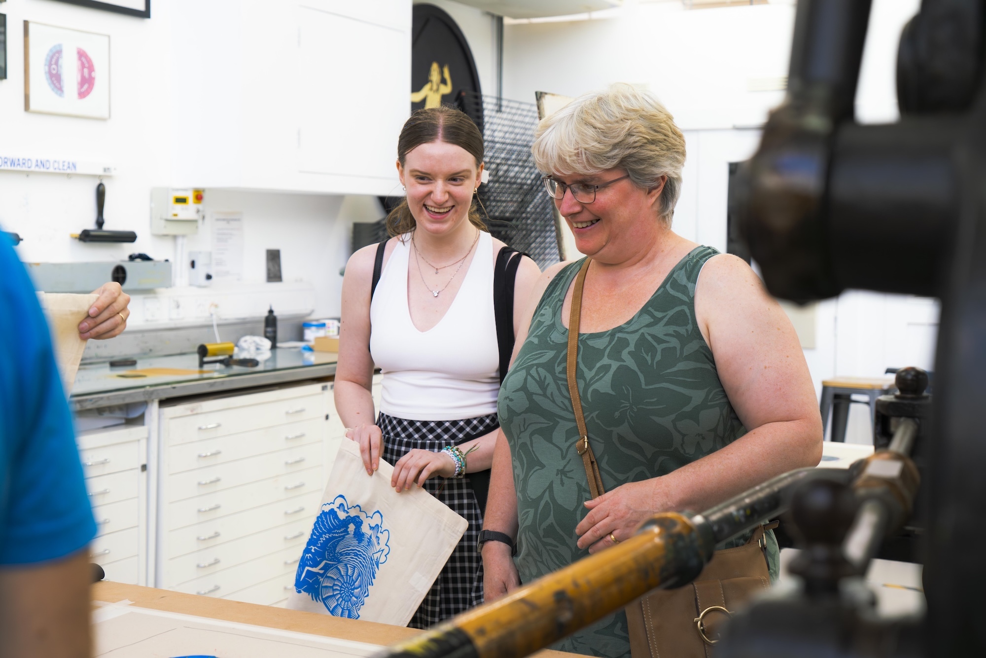 Two people standing in a studio at Arts University Bournemouth, one wearing green sleeveless top, other in white vest with patterned skirt, both smiling.