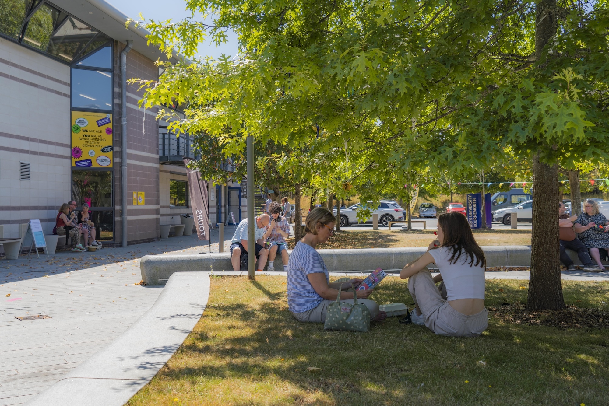 Two women sitting on grass under a large tree in an urban square, with grey buildings and parked cars visible in the background.