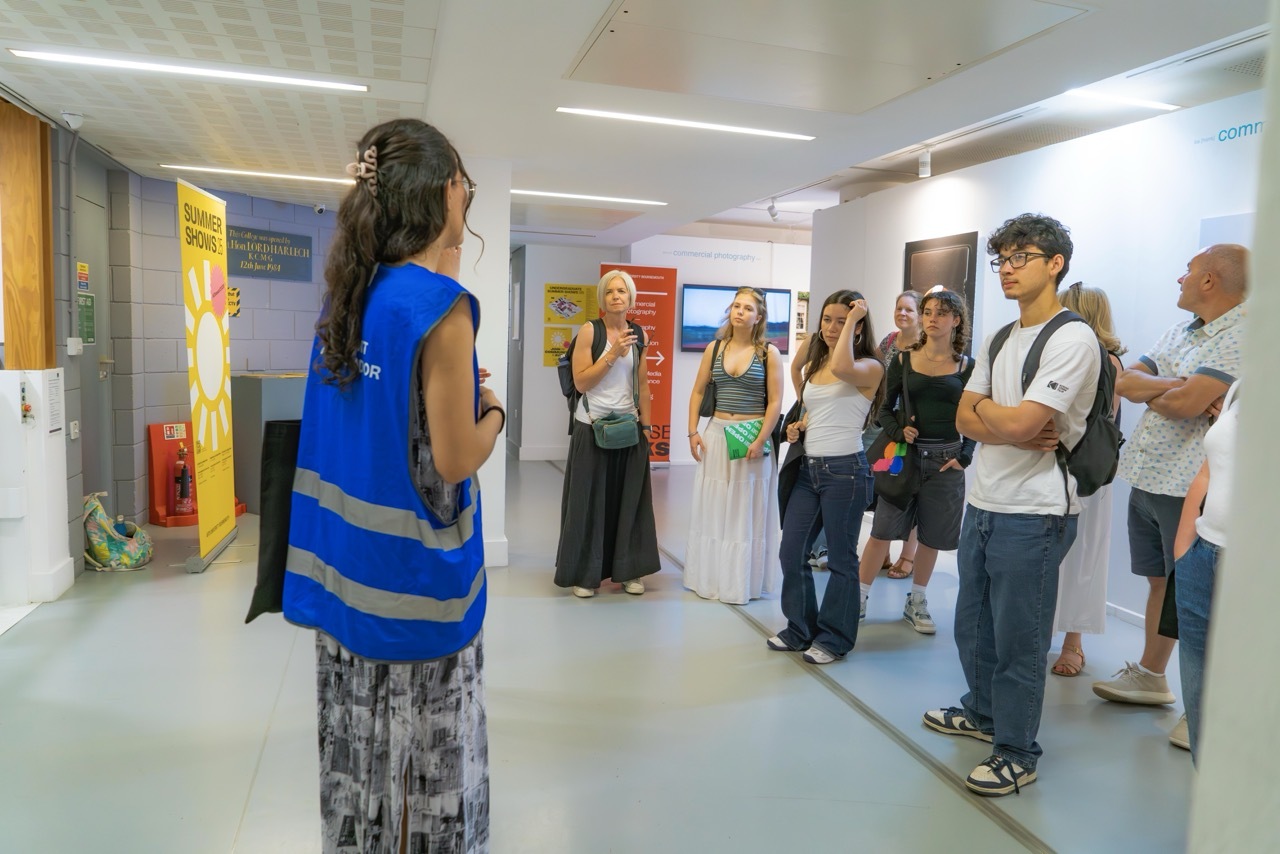 Woman in blue vest addresses group of visitors in white corridor with colourful wall displays and fluorescent lighting.