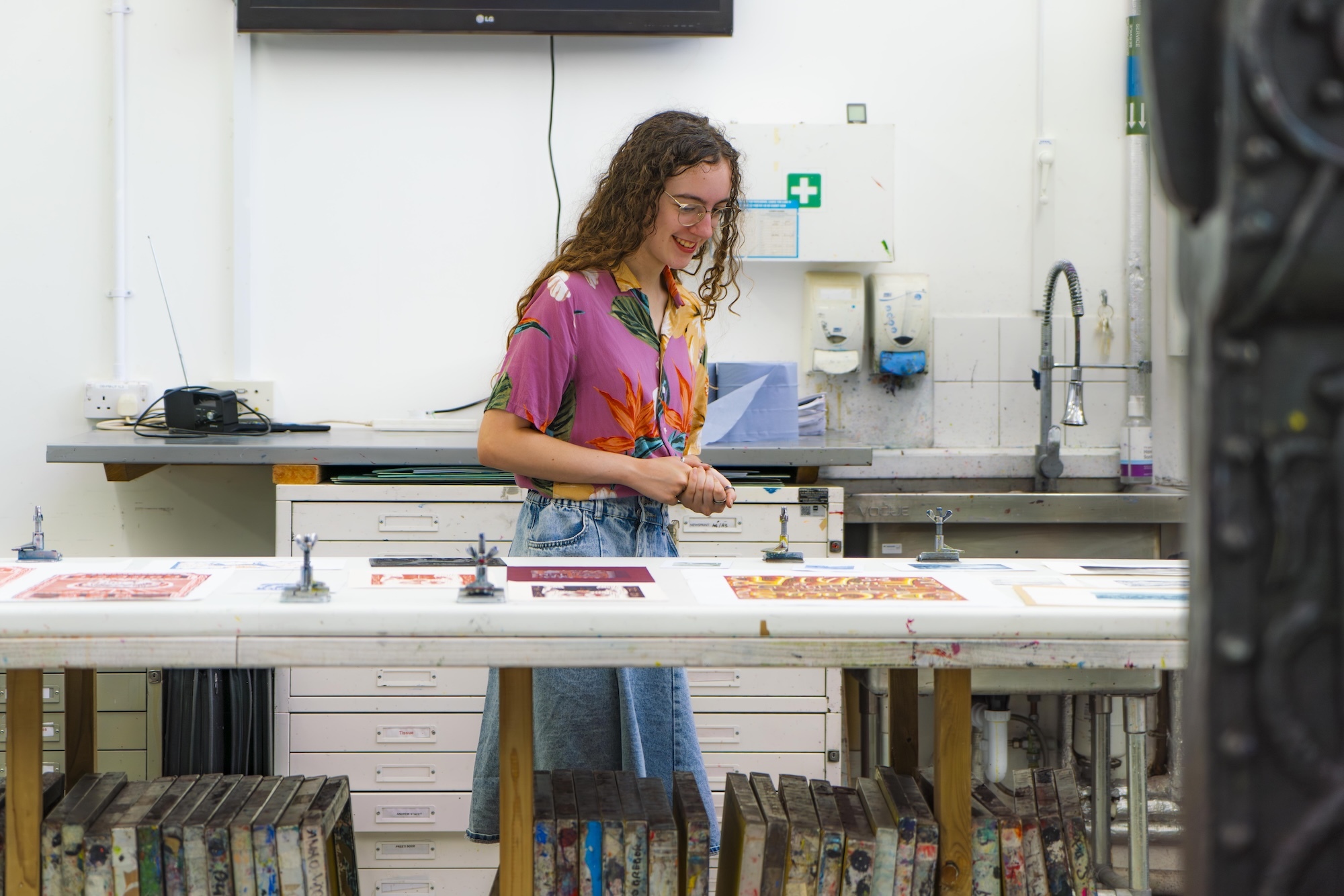 Woman with curly hair in pink top working at white table with coloured papers in bright studio space with equipment and shelving.